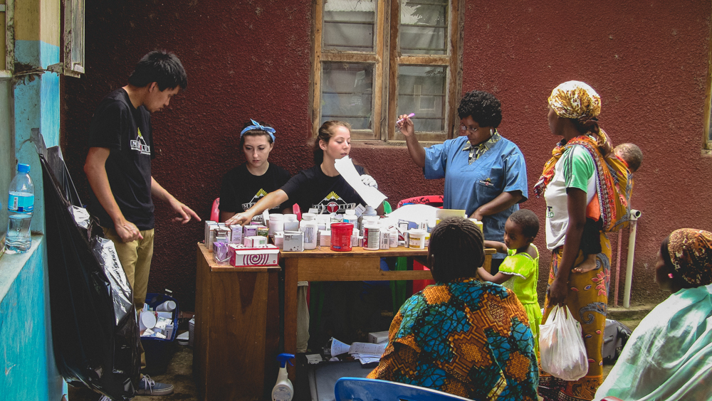 MEDLIFE students and community members gather at an outdoor mobile clinic pharmacy station against a reddish wall in Moshi, Tanzania