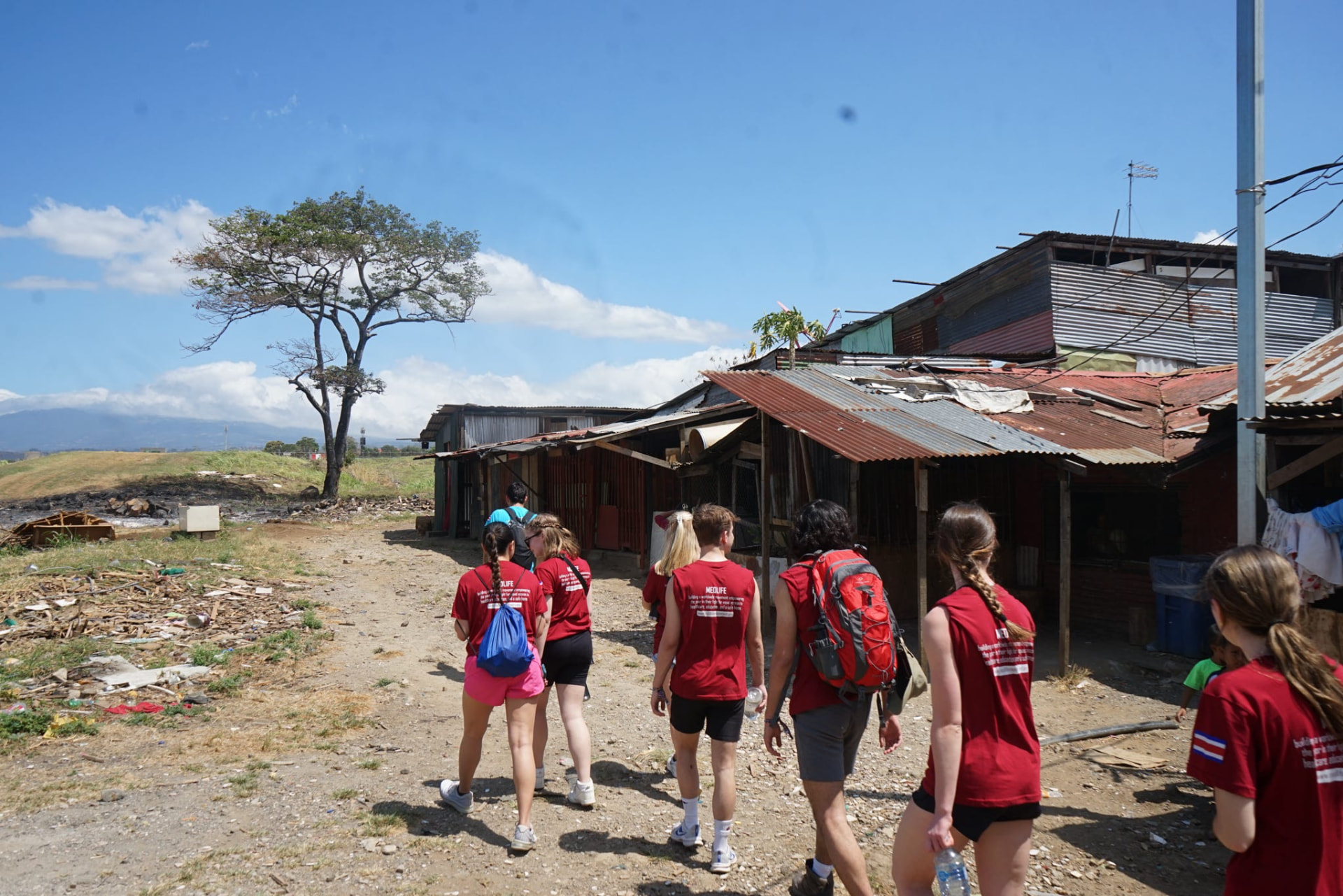 MEDLIFE students in red t-shirts walk along a dusty path past corrugated-metal dwellings in a San José, Costa Rica community