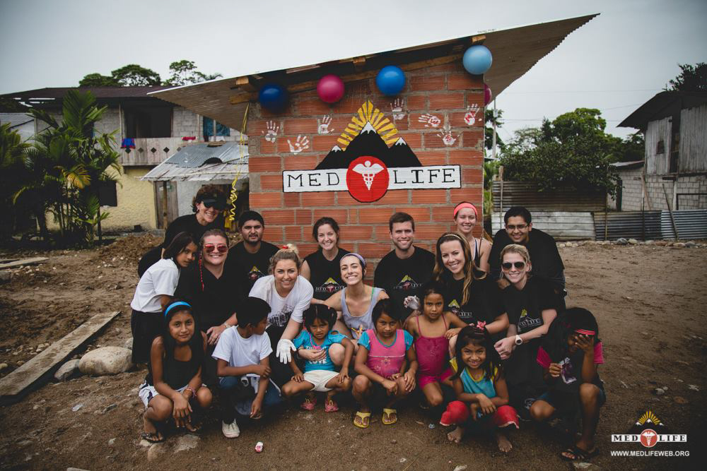 MEDLIFE students and local children pose in front of a brick community building decorated with a MEDLIFE mural and balloons near Tena, Ecuador